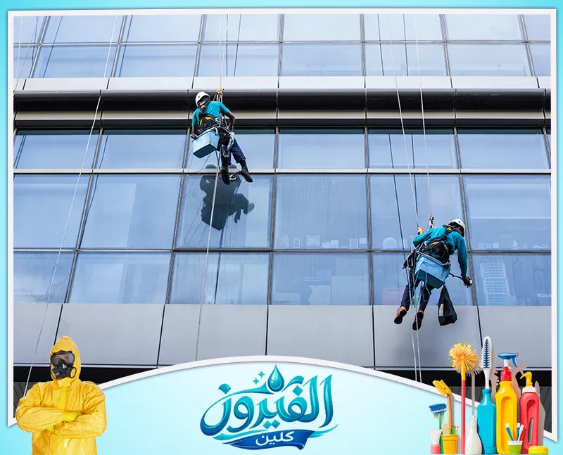 تنظيف سلالم العمائر بالدمام 0562185880 شركة الفيروز عاملان معلقان بالحبال ينظفان واجهة زجاجية ضخمة لبرج حديث، يجسدان الشجاعة والإتقان في العمل على ارتفاعات شاهقة.