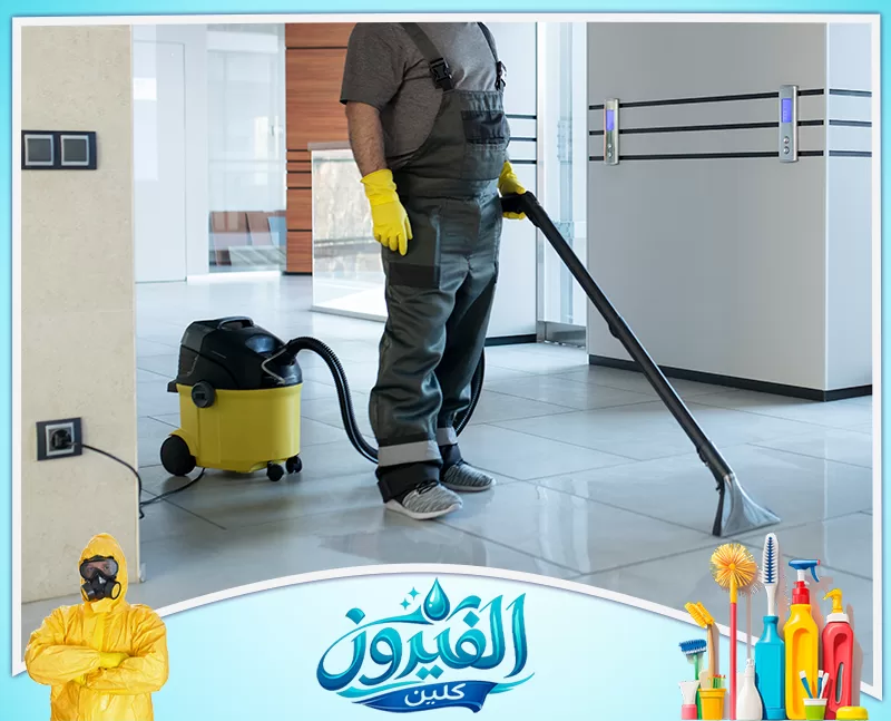 تنظيف سلالم العمائر في الجبيل 0562185880 شركة الفيروز عامل نظافة يرتدي زياً مخصصاً وقفازات صفراء، يستخدم مكنسة كهربائية صناعية لتنظيف أرضية صالة واسعة
