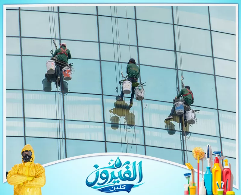 تنظيف جدران المباني من الداخل والخارج في الدمام 0562185880 شركة الفيروز ثلاثة عمال يتدلون بمهارة لتنظيف واجهة زجاجية زرقاء، يرفعون شعار الدقة والأمان في رعاية المباني الشاهقة بجدية تامة.