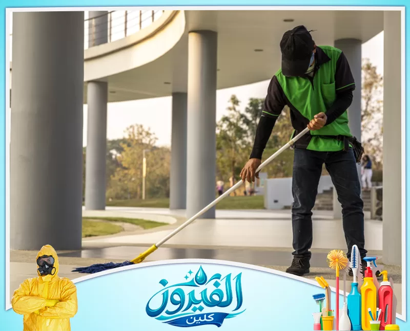 تنظيف الواجهات الزجاجية والأبواب الخارجية 0562185880 شركة الفيروز عامل نظافة يرتدي قميصاً أخضر وقبعة، يقوم بمسح الأرضية في مساحة خارجية واسعة تحت سقف مدعوم بأعمدة.