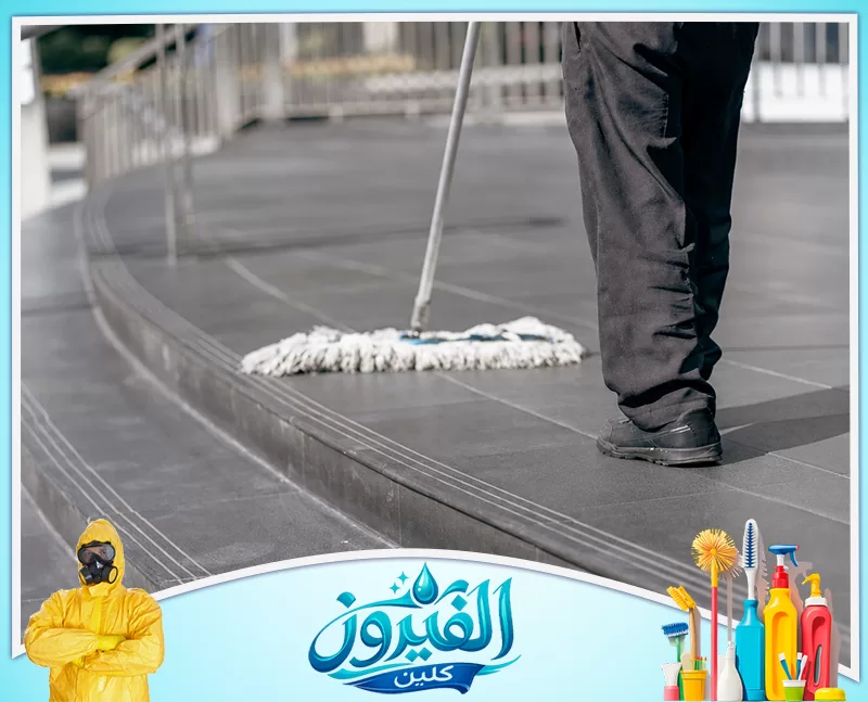 تنظيف السلالم والممرات في العمائر بالدمام 0562185880 شركة الفيروز عامل نظافة يمسح أرضية رمادية بجد واجتهاد، يظهر أسفل جسده وهو ممسك بالممسحة فوق بلاط نظيف ولامع.