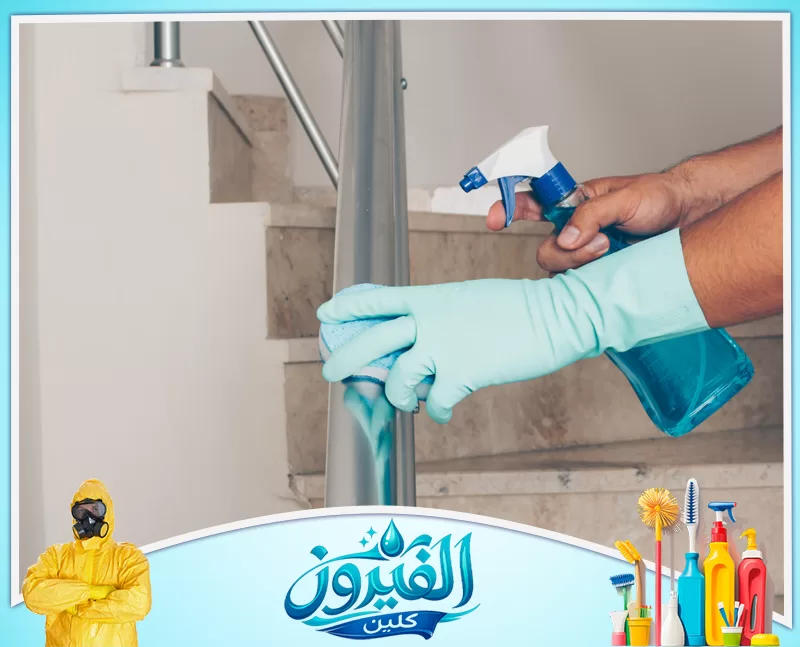 تنظيف المداخل واللوبي بالظهران 0562185880 شركة الفيروز شخص يرتدي قفازات خضراء ينظف درابزين سلم معدني باستخدام إسفنجة وزجاجة رش بها سائل أزرق.