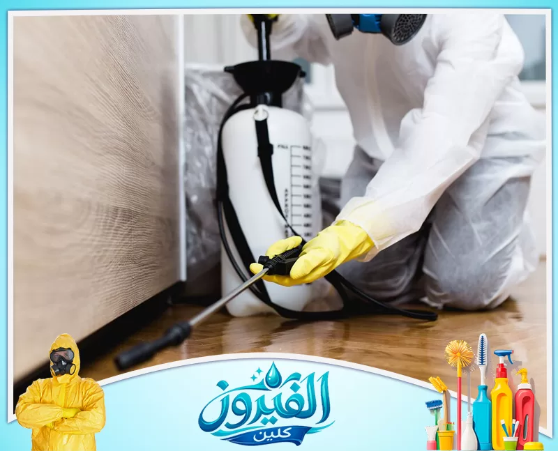 مكافحة الصراصير بالظهران 0562185880 شركة الفيروز خبير يرتدي ملابس حماية يرش مبيدات أسفل خزانة خشبية، لحماية المنزل من الحشرات الضارة.